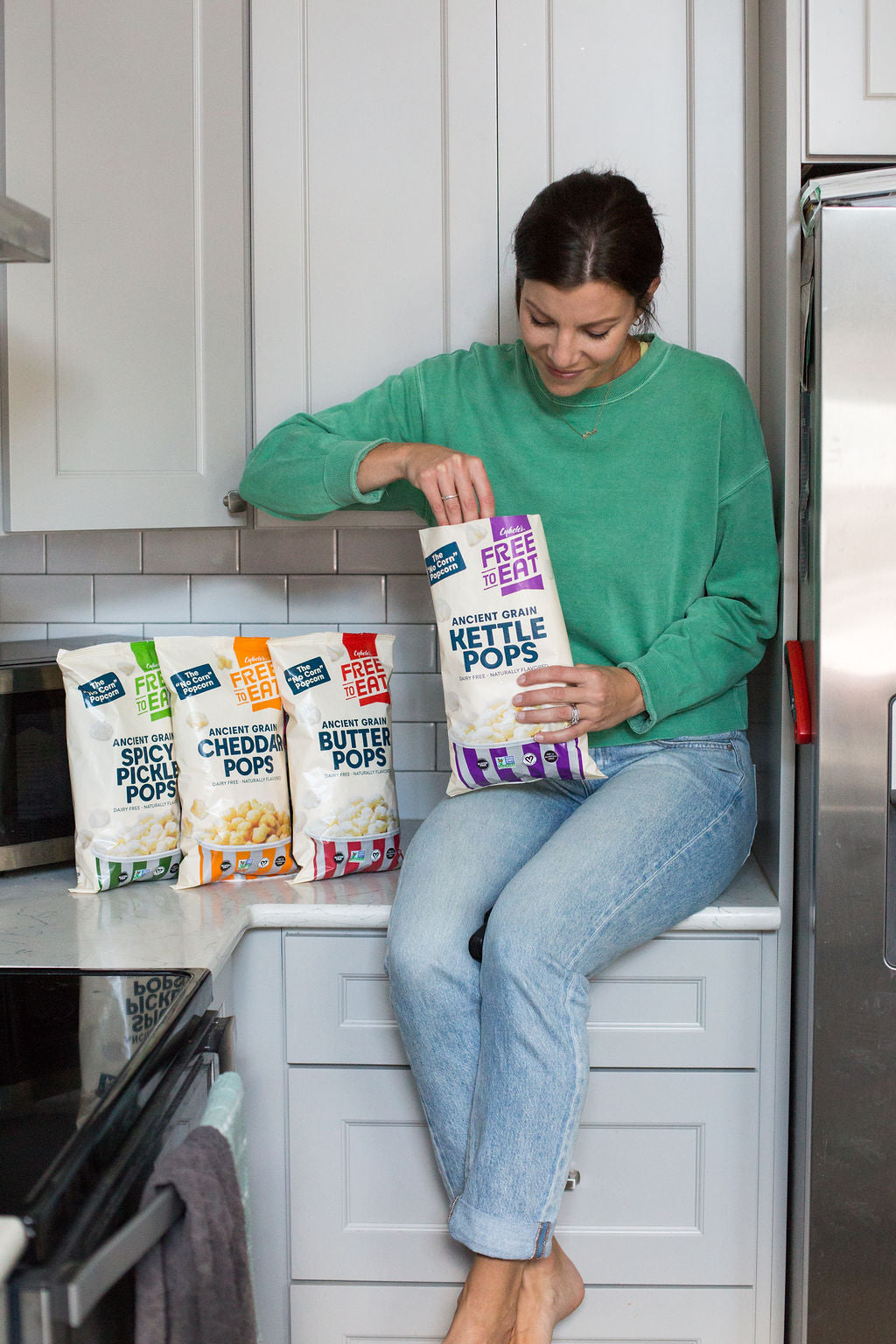 Woman wearing green sweatshirt and jeans sitting on counter with 4 bags of Ancient Grain Pops