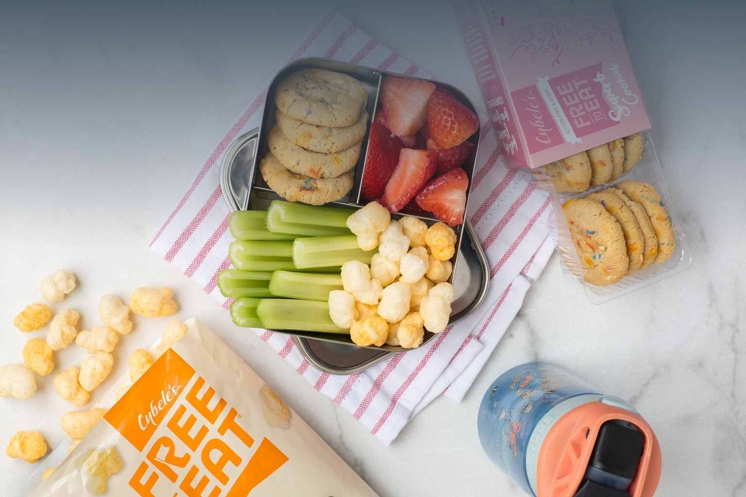 kids bento box on counter with Bag of Cybeles Cheddar Ancient Grain Pops, box of Confetti Cookies and Water bottle. Bento is filled with celery, ancient grain Pops, confetti cookies and strawberries