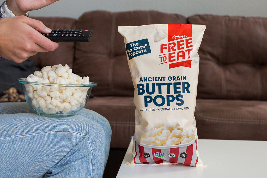 Person watching TV with a remote in hand and bowl of Butter Pops on lap. Bag of Butter Ancient Grain Pops are on the coffee table and brown leather couch is shown in background