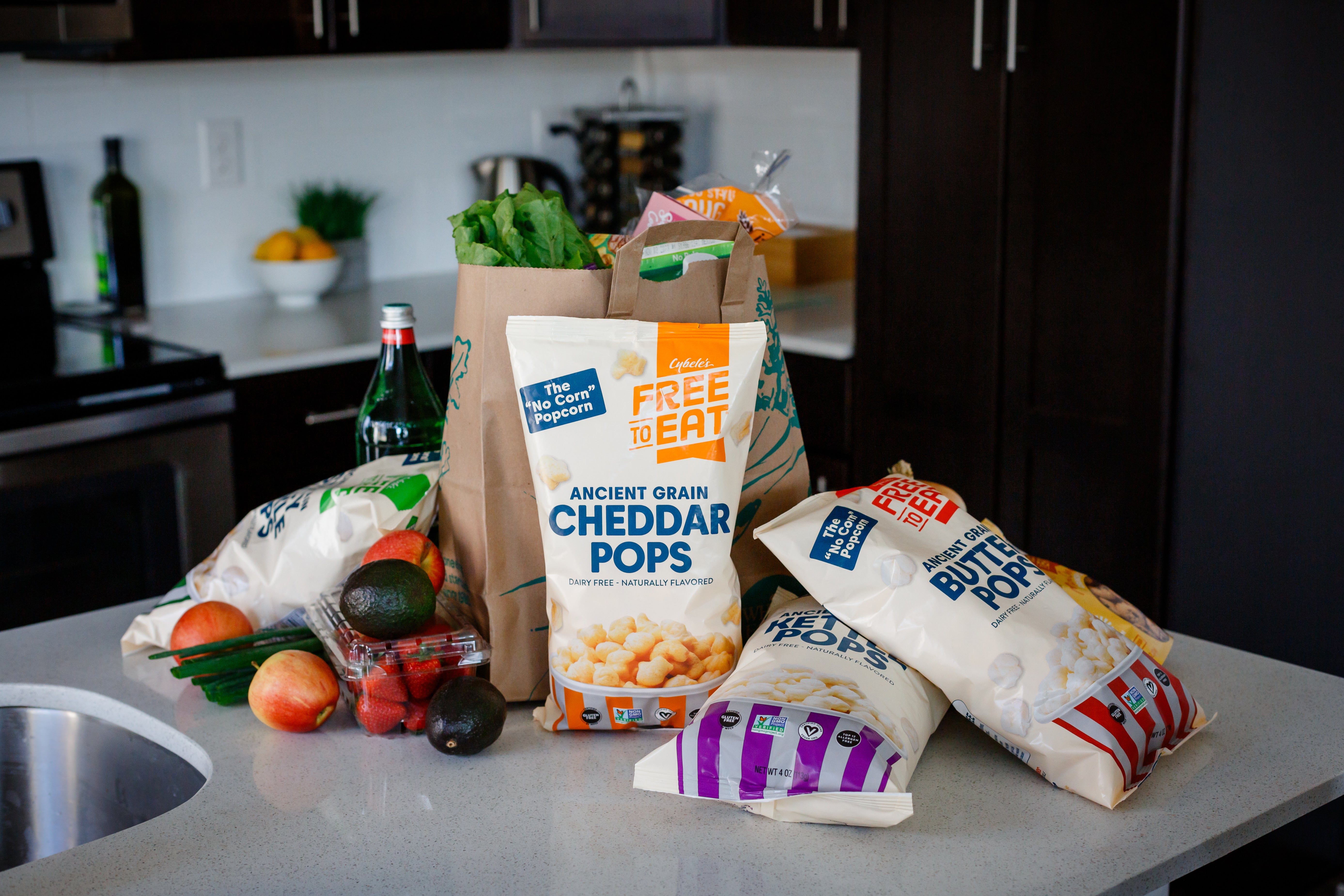 Pile of ancient grain pops bags on kitchen counter with paper sack of groceries and vegetables