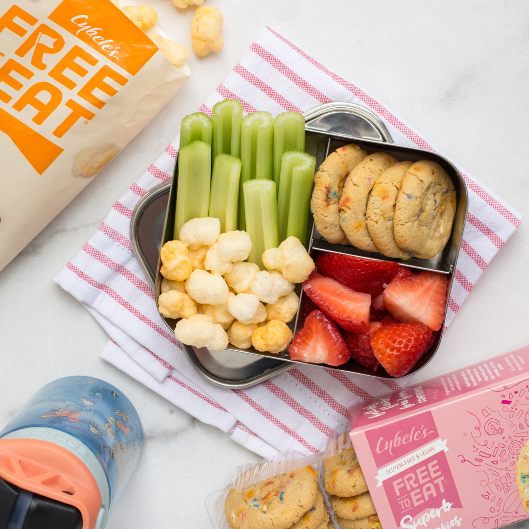 kids bento box on counter with Bag of Cybeles Cheddar Ancient Grain Pops, box of Confetti Cookies and Water bottle. Bento is filled with celery, ancient grain Pops, confetti cookies and strawberries